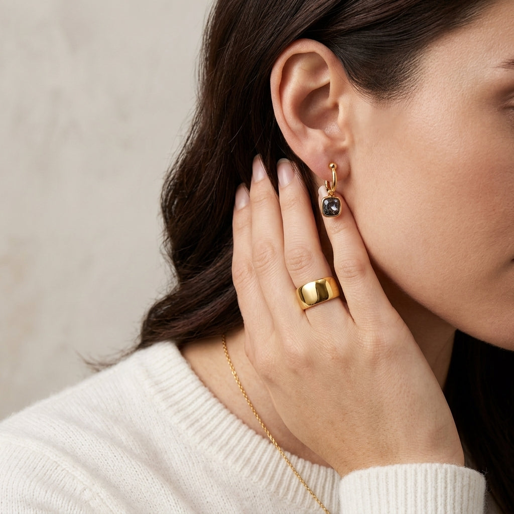 A close-up lifestyle shot of a woman wearing a delicate gold huggie hoop earring with a small black stone, along with a gold ring and necklace.