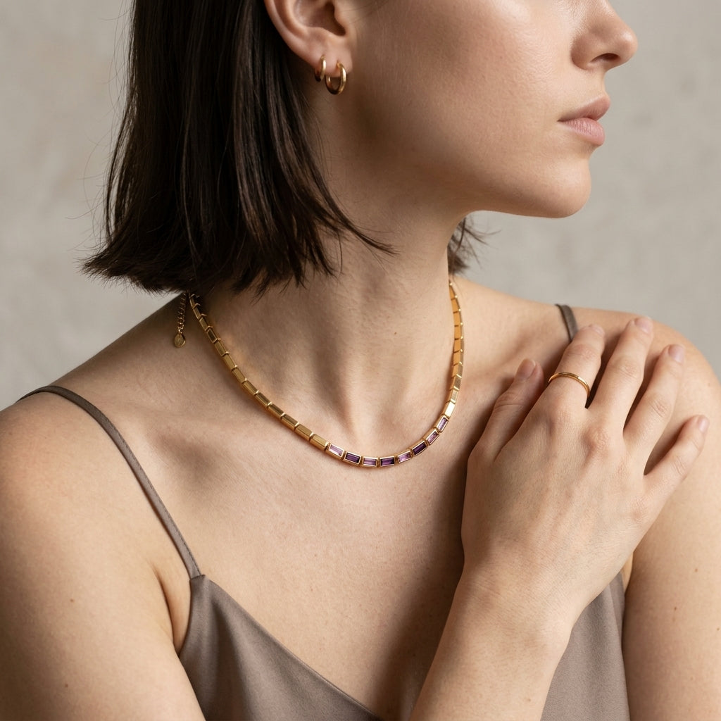 A close-up lifestyle shot of a woman wearing a gold beaded necklace with small purple gemstone accents, styled with a taupe camisole against a neutral textured background.