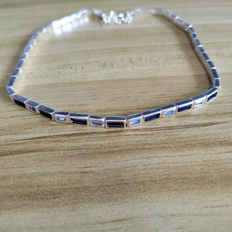 A silver tennis-style necklace with rectangular blue and black stones, displayed on a light wood grain surface.