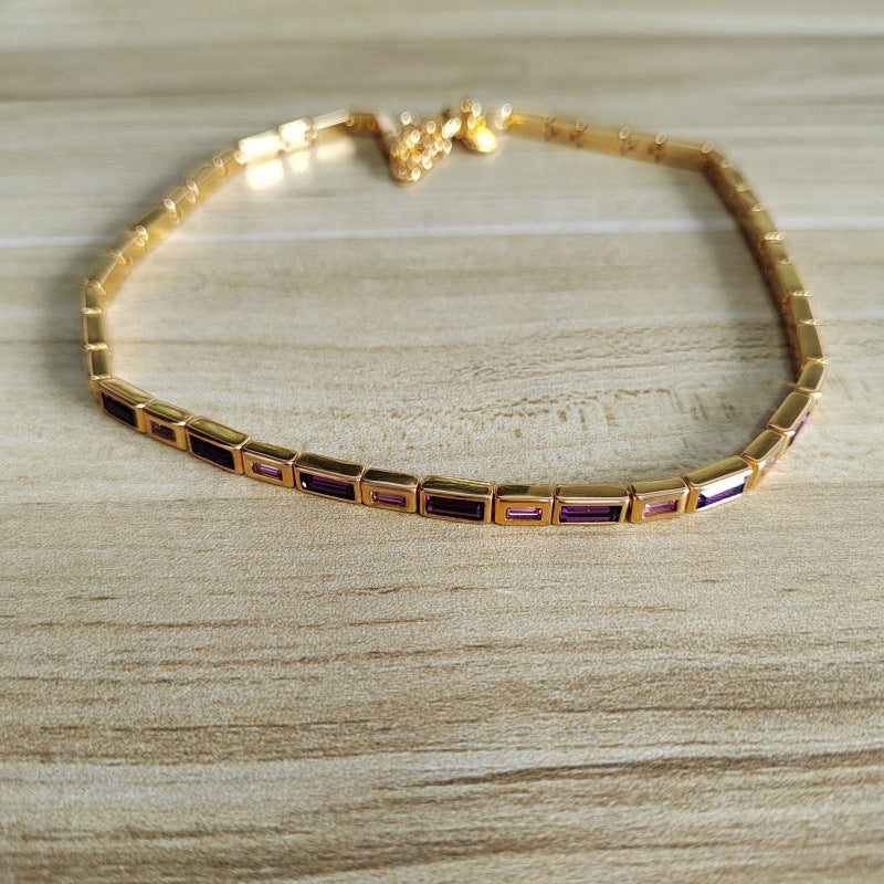 A gold-plated tennis bracelet with rectangular bezel-set purple and black stones, displayed on a light wood grain surface.