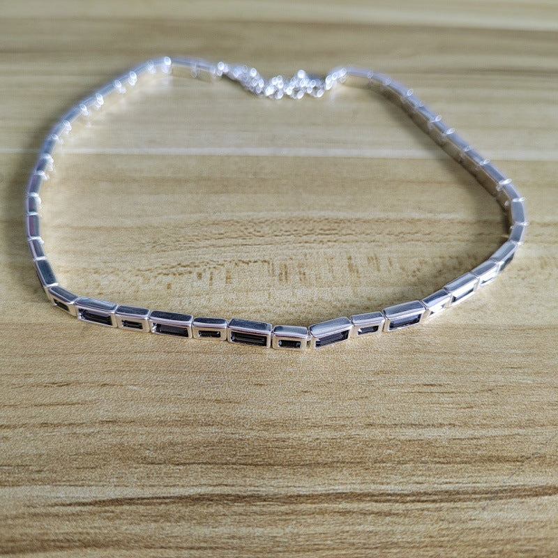 A silver tennis-style necklace with rectangular clear stones, displayed on a light wood grain surface.