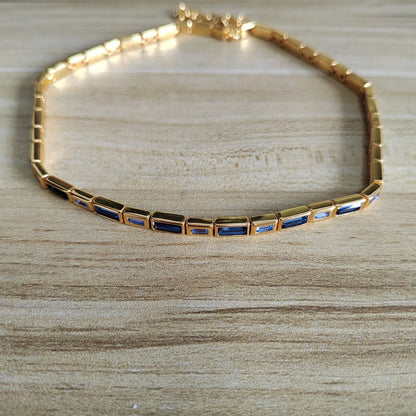 A gold tennis-style bracelet with rectangular blue inlay stones, displayed on a light wood surface.