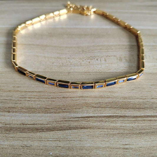 A gold tennis-style bracelet with rectangular blue inlay stones, displayed on a light wood surface.