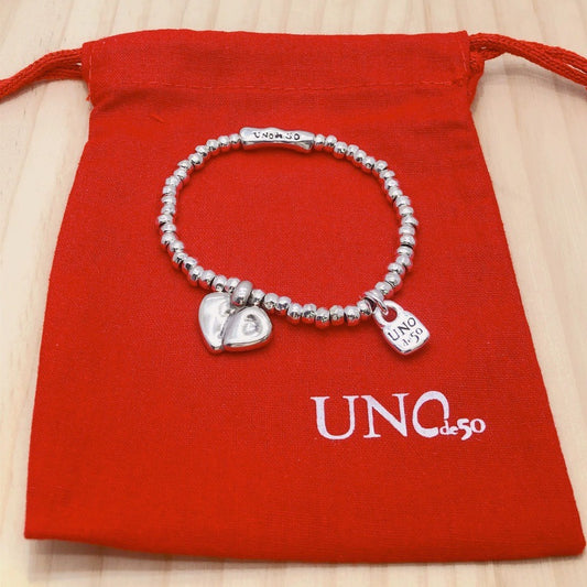 A silver beaded bracelet with heart and padlock charms, displayed on a red drawstring pouch with the brand name 'UNO de 50' printed on it, set against a light wood background.
