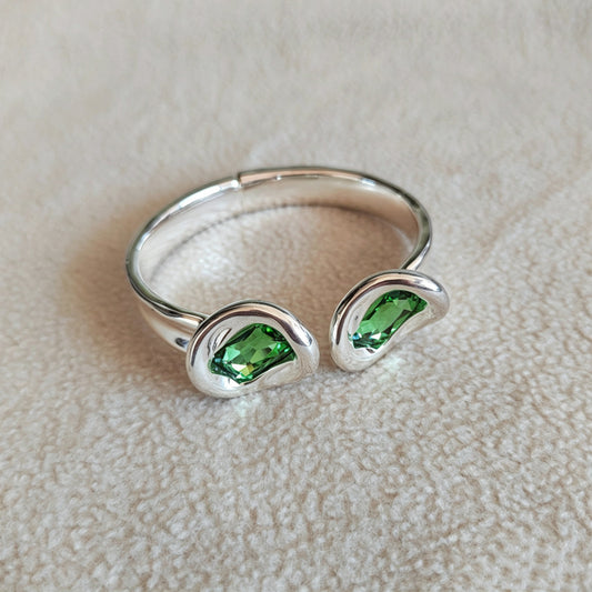 A silver open cuff ring featuring two wavy, open-ended sides, each set with a small, faceted green gemstone, resting on a textured beige fabric surface.