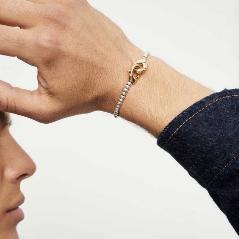 A close-up of a person's wrist wearing a gold bracelet with a dolphin design and a silver beaded chain segment, shown against a plain light background with part of the person's face and denim sleeve visible.