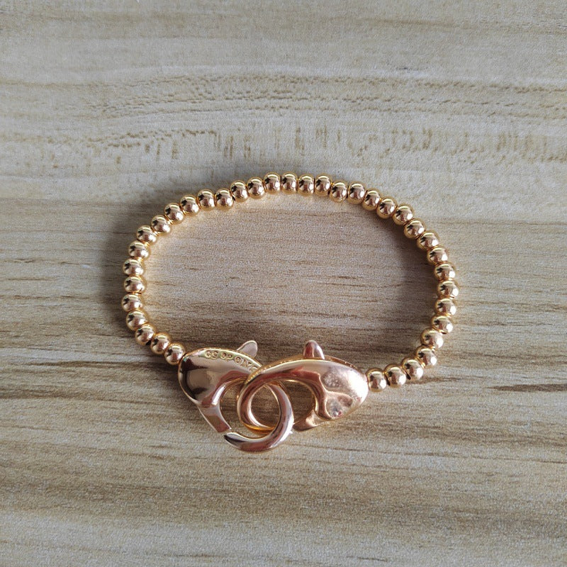 A rose gold beaded bracelet with a lobster clasp featuring a small fish charm, displayed on a light wood grain surface.