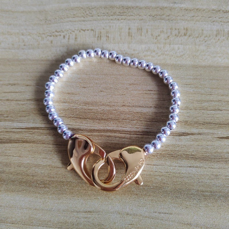 A beaded bracelet with a silver-toned beaded band and a rose gold lobster clasp featuring two interlocking fish shapes, displayed on a light wood background.