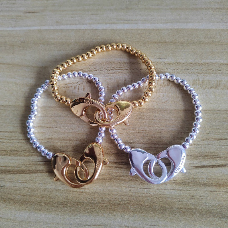 Three beaded bracelet-style rings with fish-shaped clasps, shown in rose gold, yellow gold, and silver tones on a light wood surface.
