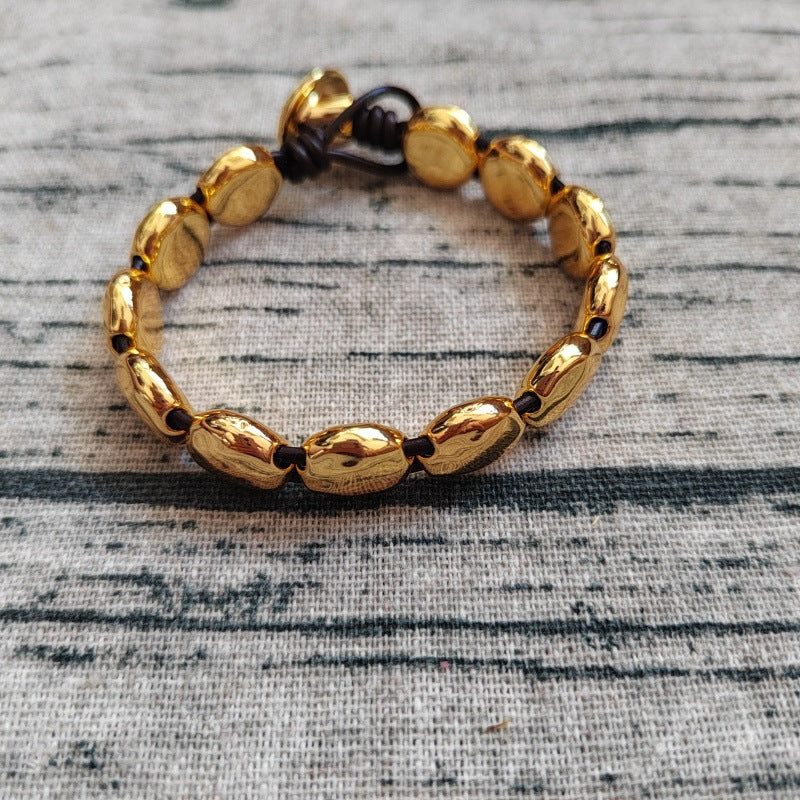 A gold-toned beaded bracelet with polished oval beads and a dark cord closure, photographed on a textured gray fabric background.