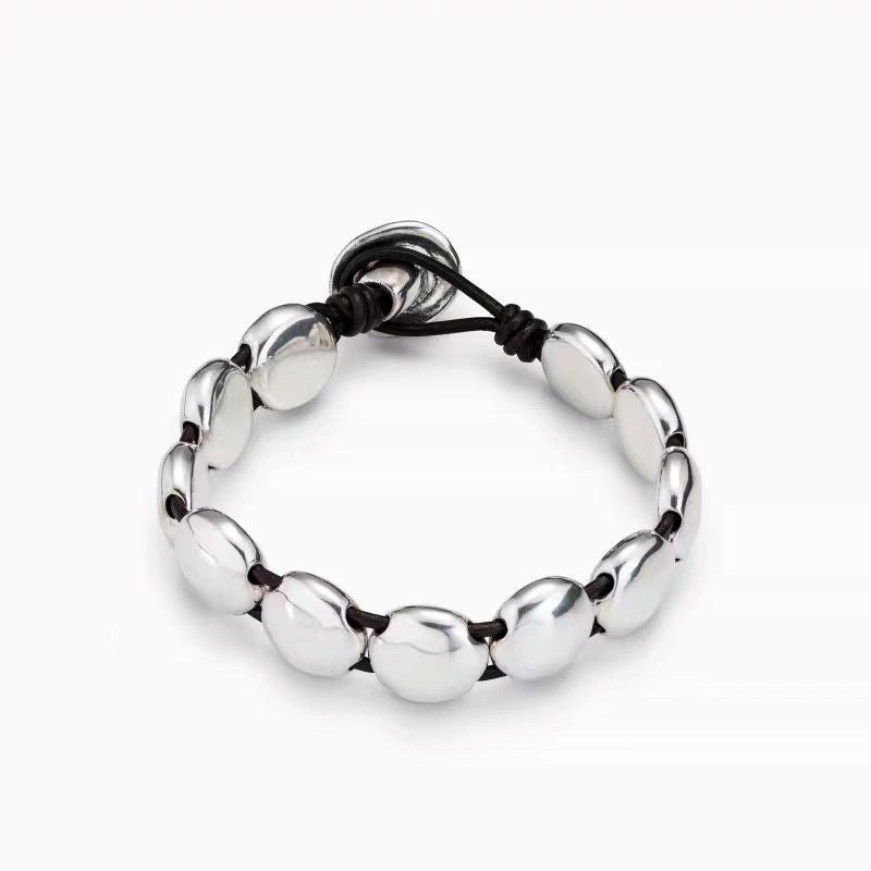 A silver-toned beaded bracelet with smooth, polished oval beads separated by thin black cords, featuring a matching silver disc charm on the adjustable cord, shown on a light gray background.
