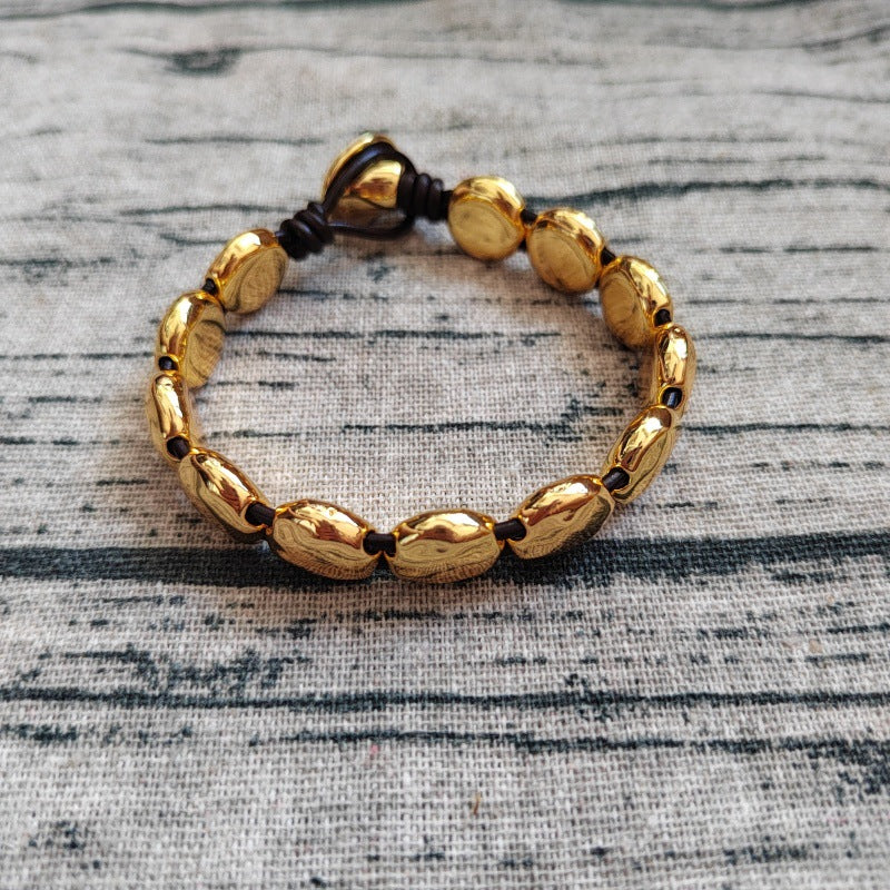 A gold-toned beaded bracelet with polished oval beads and a dark brown adjustable cord closure, displayed on a textured gray fabric background.