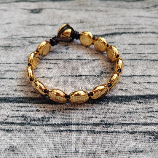 A gold-toned beaded bracelet with polished oval beads and a dark brown adjustable cord closure, displayed on a textured gray fabric background.
