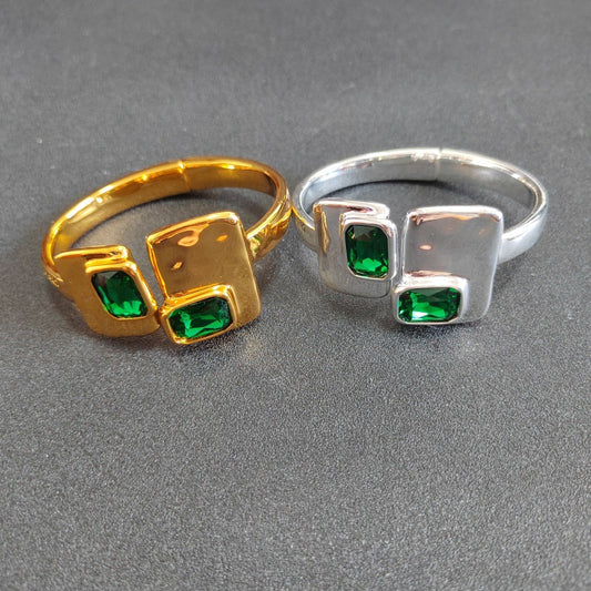 Two modern square ring designs shown side-by-side on a textured gray background. The left ring is gold-plated with two green rectangular gemstones, and the right ring is silver-plated with two matching green rectangular gemstones.