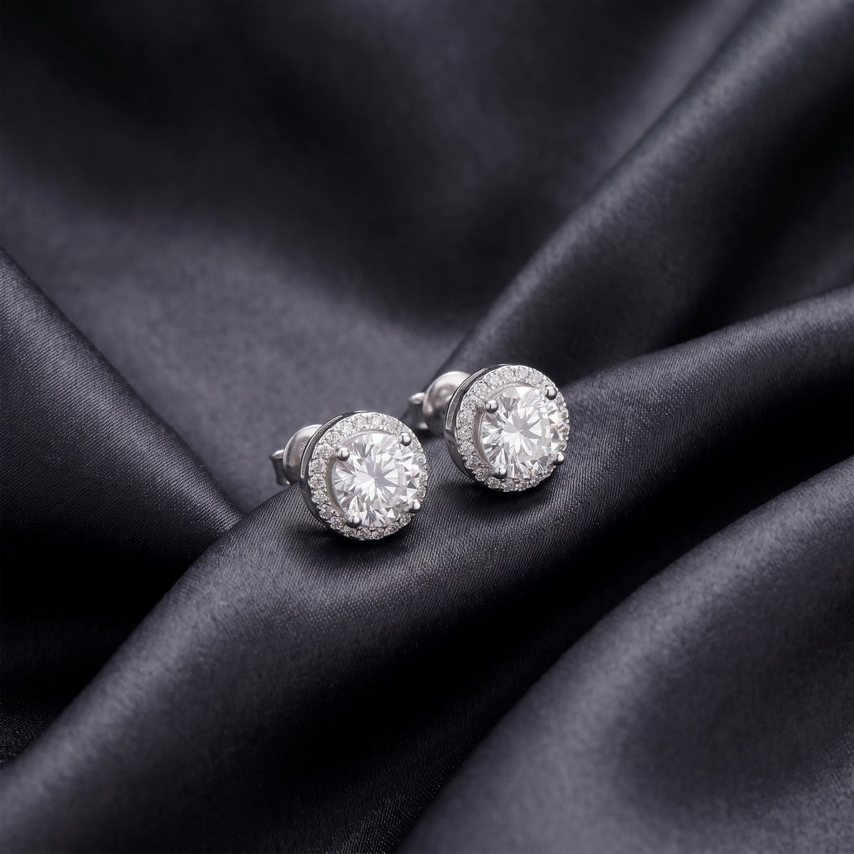 Pair of round halo lab grown diamond stud earrings placed at an angle on dark black silk
