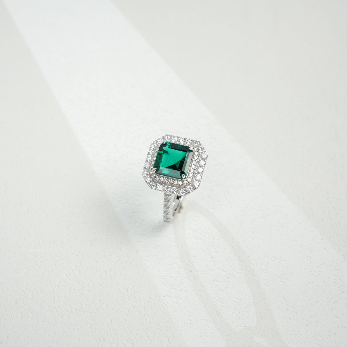 Three-quarter studio product view of a classic step-cut hydro-thermal emerald double halo engagement ring resting on a white surface.