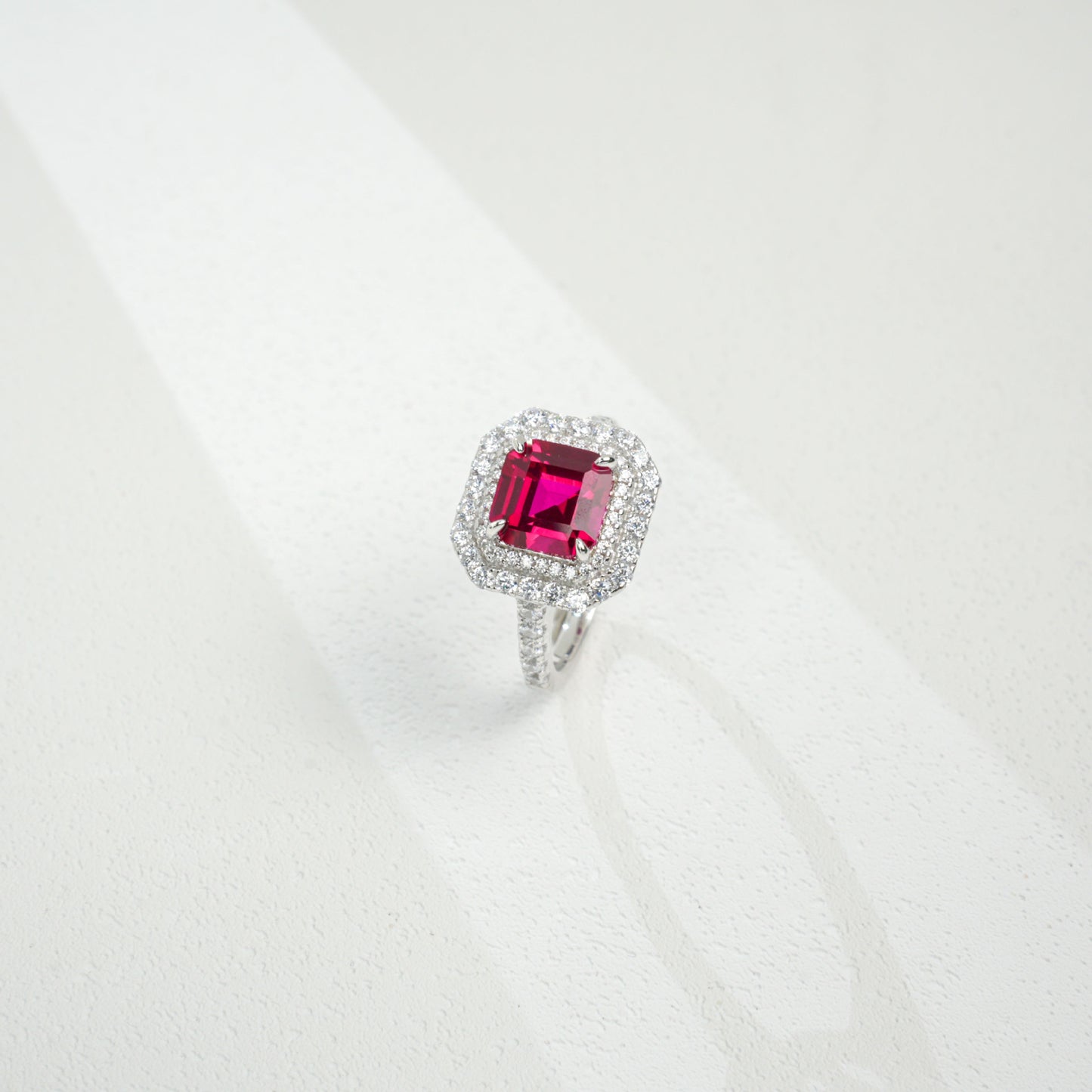 Three-quarter studio product view of a classic step-cut ruby double halo engagement ring resting on a white silk-like surface.