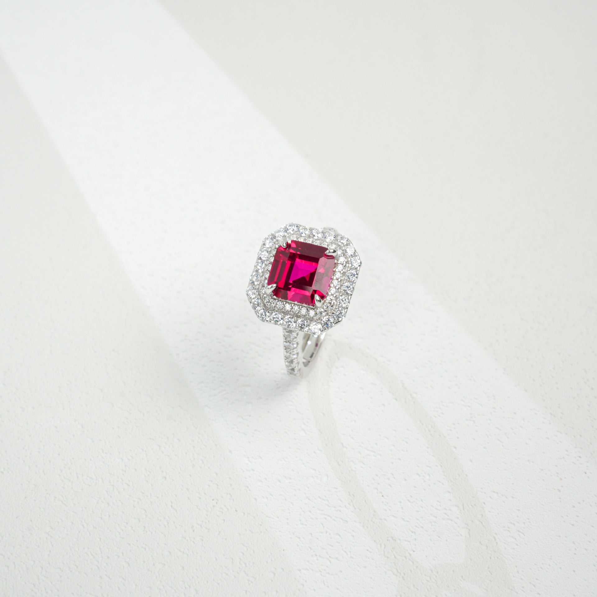 Three-quarter studio product view of a classic step-cut ruby double halo engagement ring resting on a white silk-like surface.
