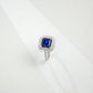 Three-quarter studio product view of a classic step-cut sapphire double halo engagement ring resting on a white silk-like surface.