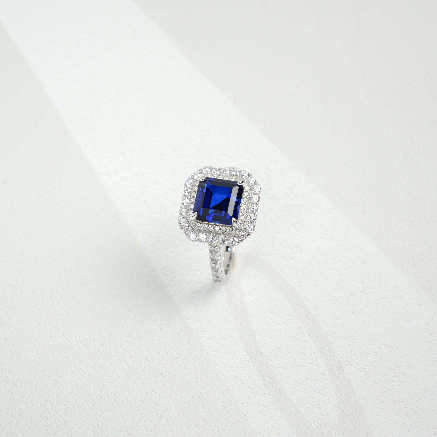Three-quarter studio product view of a classic step-cut sapphire double halo engagement ring resting on a white silk-like surface.