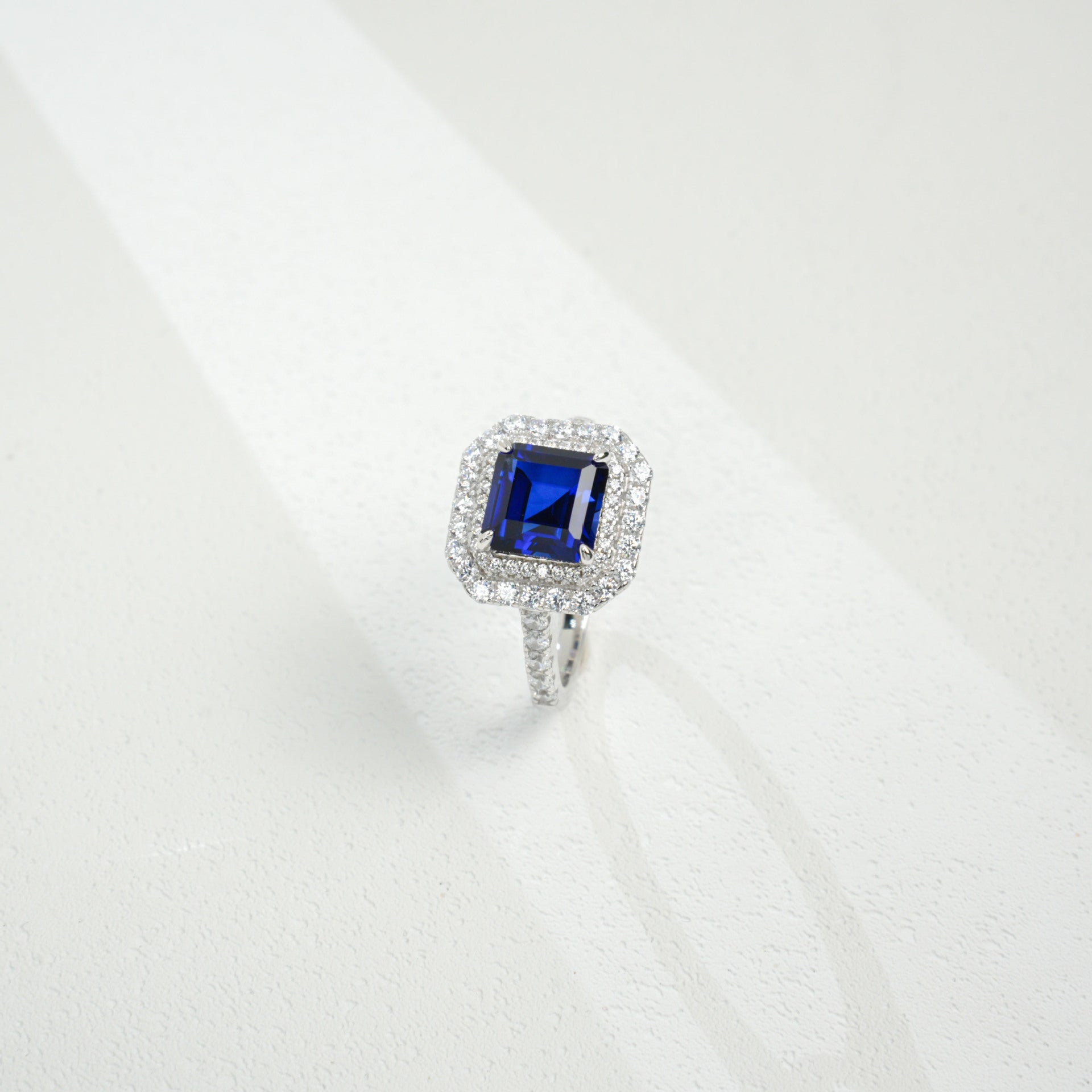 Three-quarter studio product view of a classic step-cut sapphire double halo engagement ring resting on a white silk-like surface.