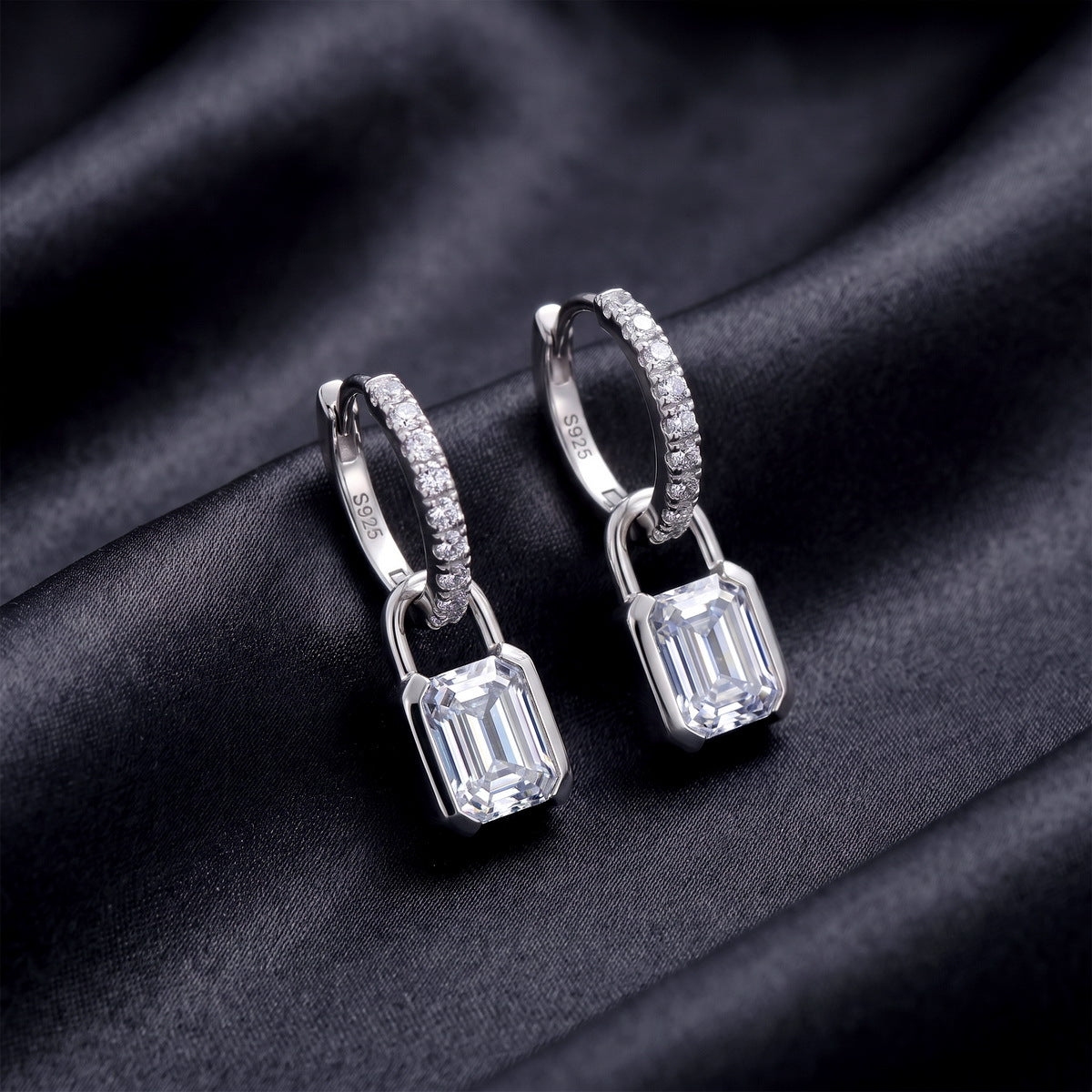Pair of emerald cut lab grown diamond padlock dangle huggie earrings resting on dark black silk