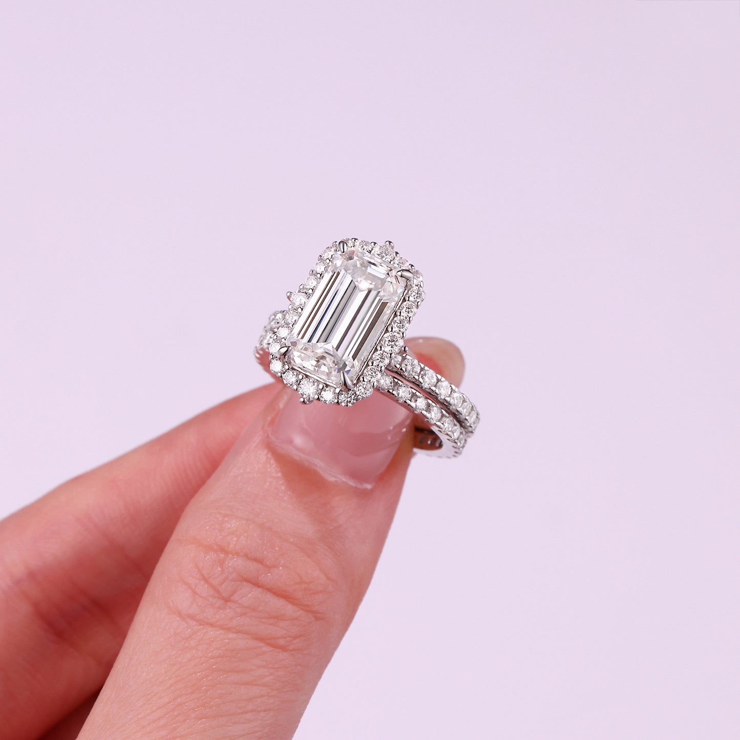 Emerald cut moissanite halo ring with matching pave band held in fingers against purple background.