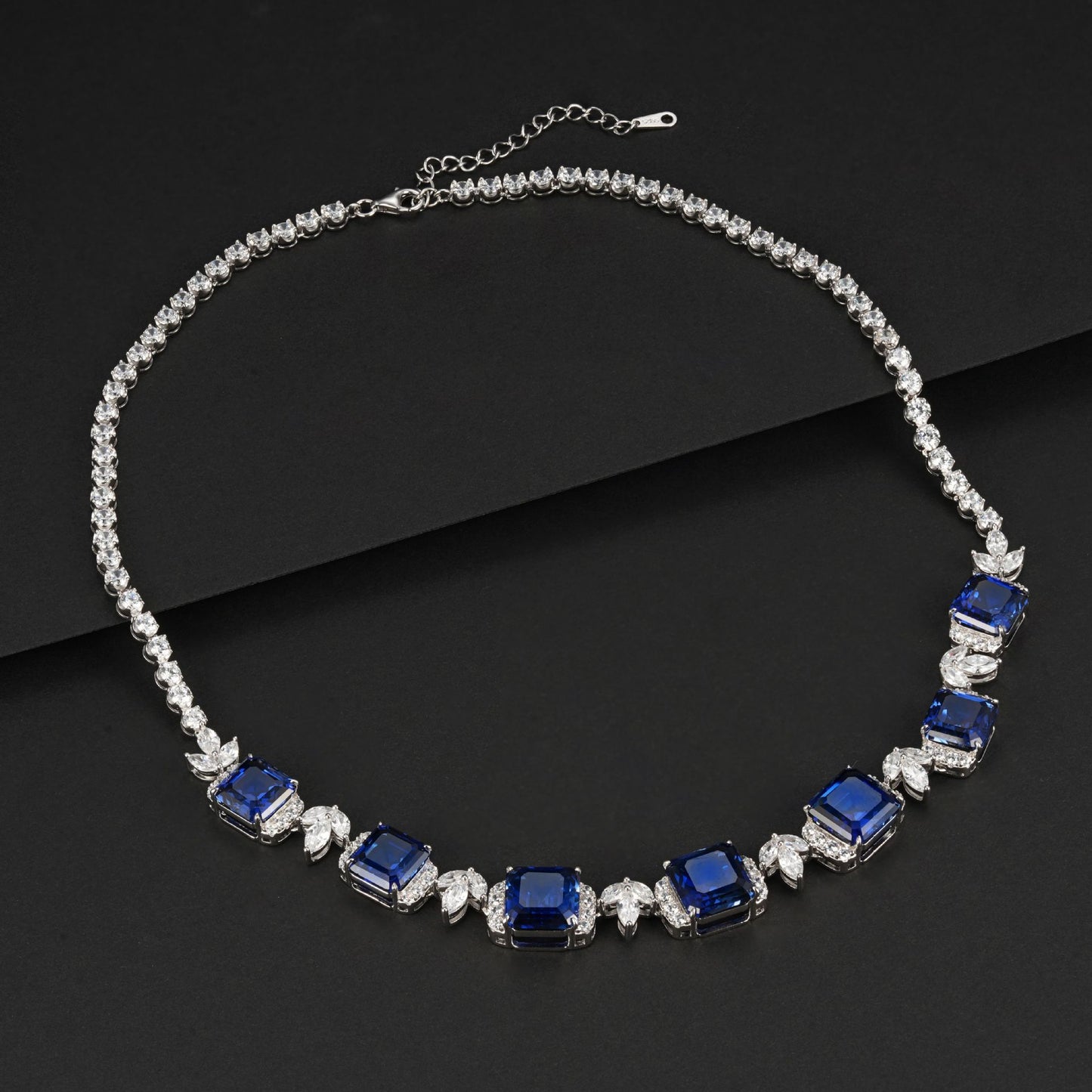 Overhead dark flat lay view of a fine jewelry blue sapphire gemstone collar necklace showcasing flexible linked prongs.