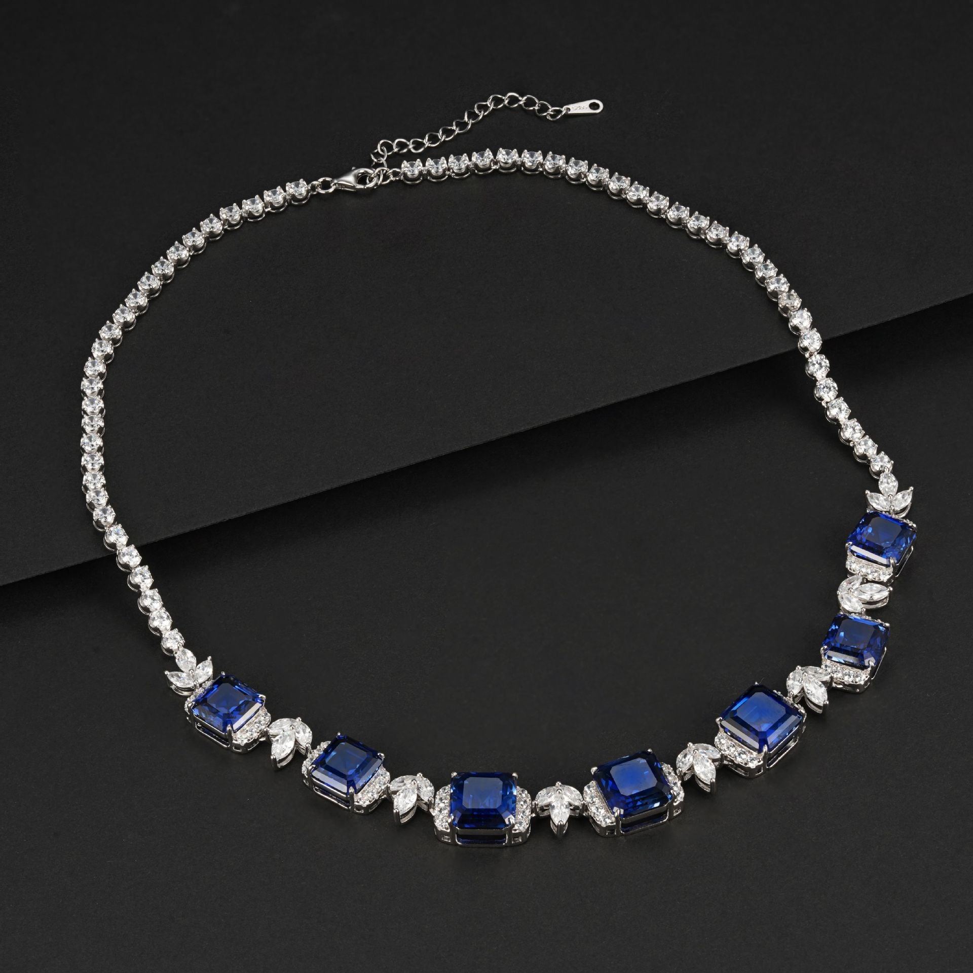 Overhead dark flat lay view of a fine jewelry blue sapphire gemstone collar necklace showcasing flexible linked prongs.