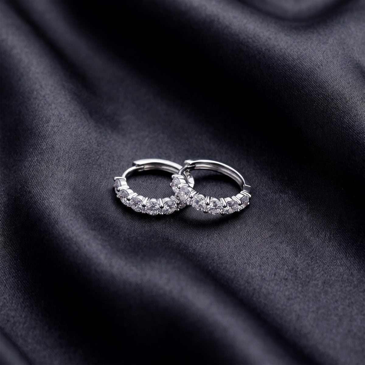 Pair of five stone round brilliant lab grown diamond huggie hoop earrings resting flat on dark black silk