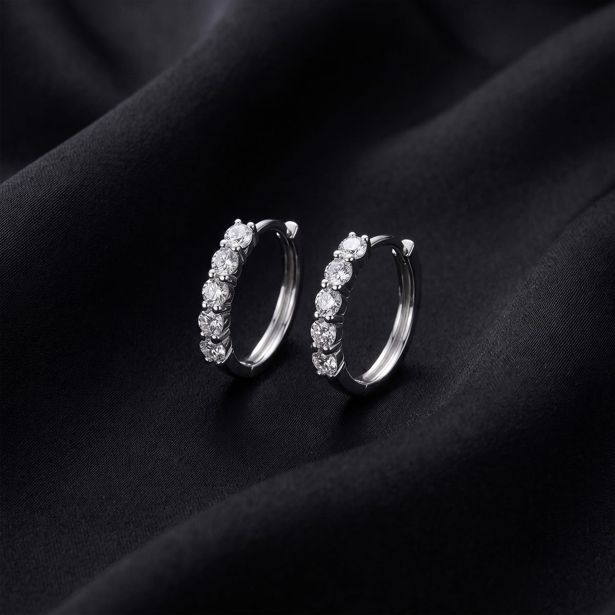 Pair of five stone round cut lab grown diamond huggie hoop earrings standing upright on dark black silk