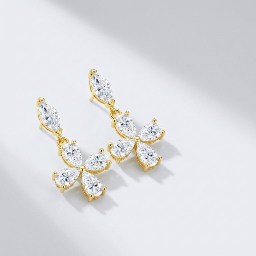 Pair of gold marquise and pear cut moissanite floral cross dangle earrings resting on a light gray surface.