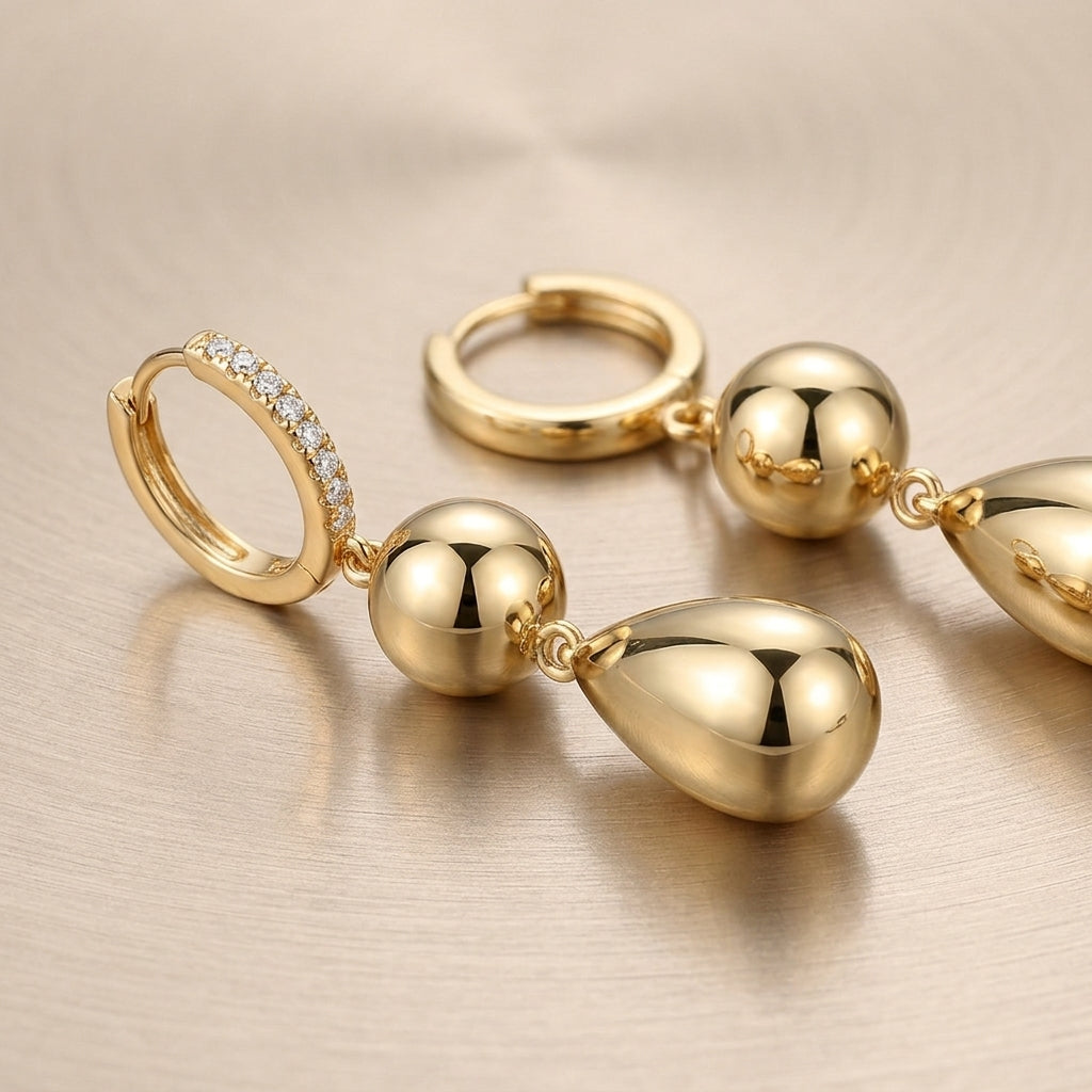 Gold tiered sphere and teardrop moissanite drop earrings lying flat on a textured surface.