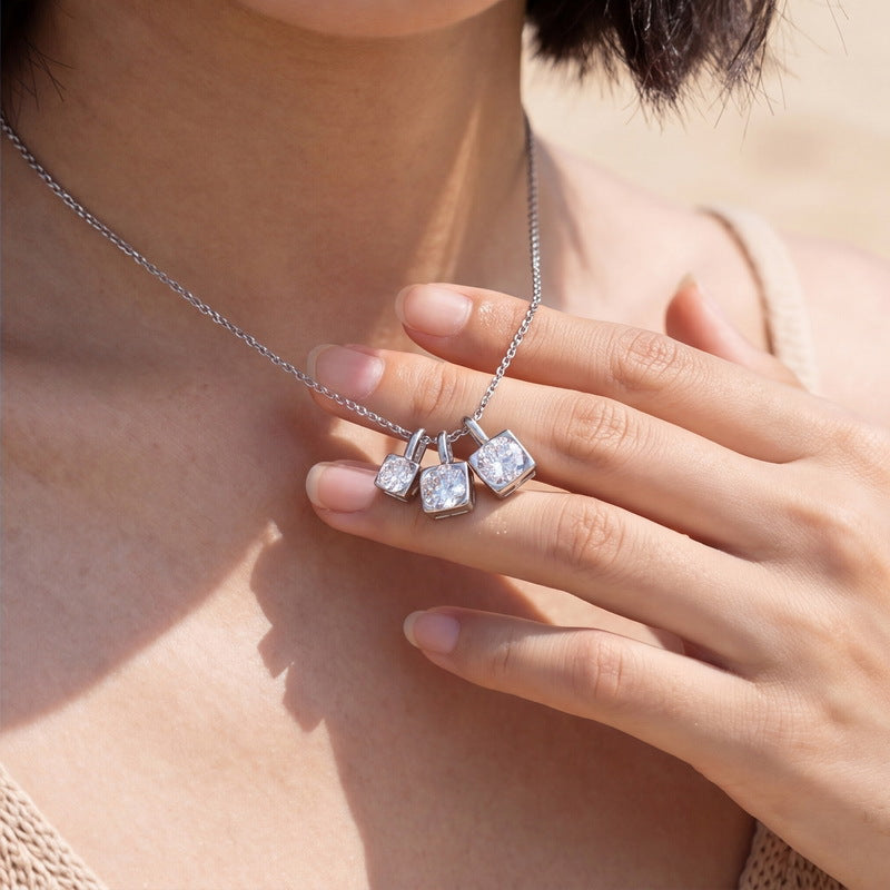 Close-up shot of model hand touching graduated moissanite trio square drop pendant by the beach.