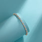 A gold tennis bracelet with clear gemstones, displayed on a light blue cylindrical stand against a matching light blue background.