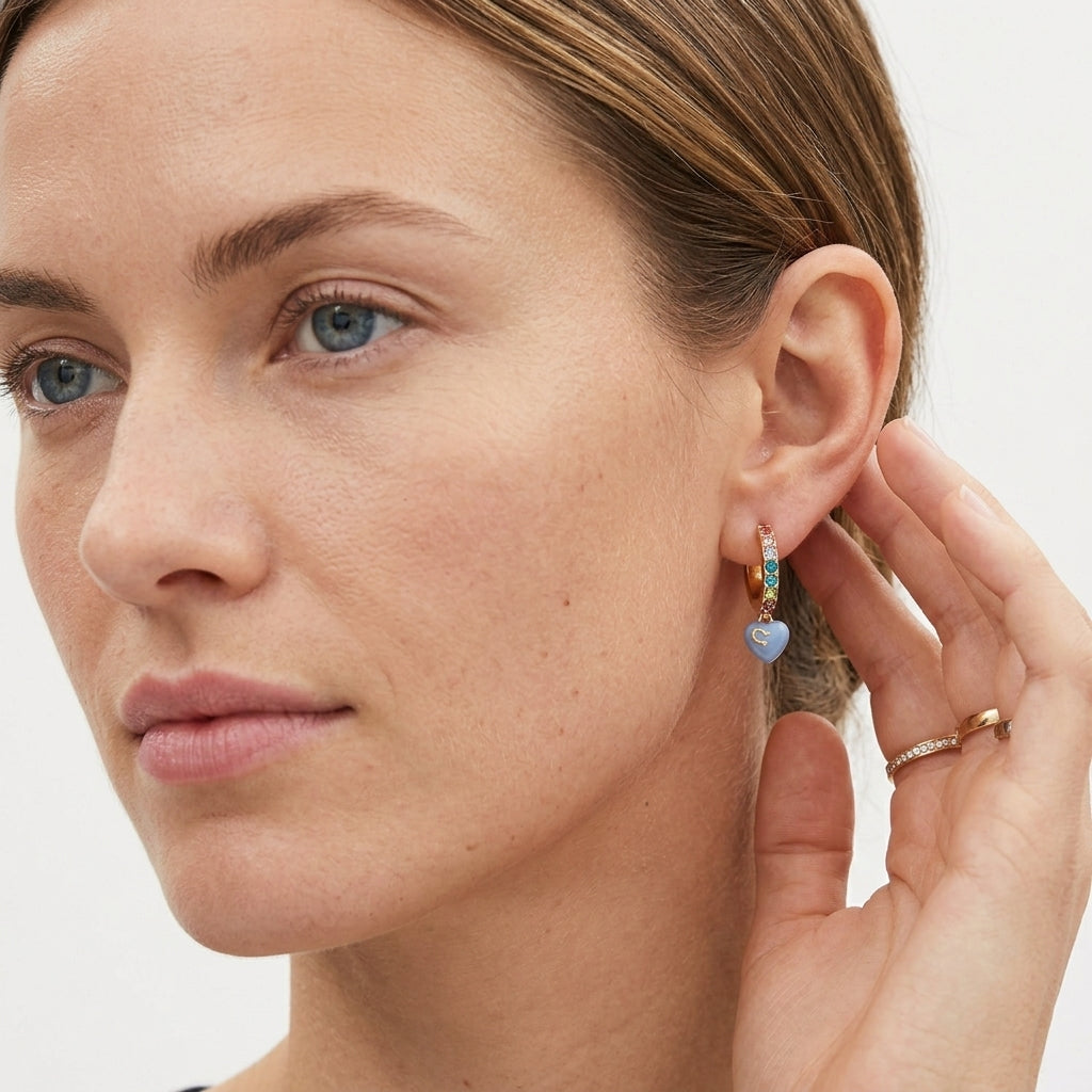 Close-up of a woman wearing a small gold huggie hoop earring with a light blue heart charm and a colorful rainbow accent, while touching her ear with her hand.