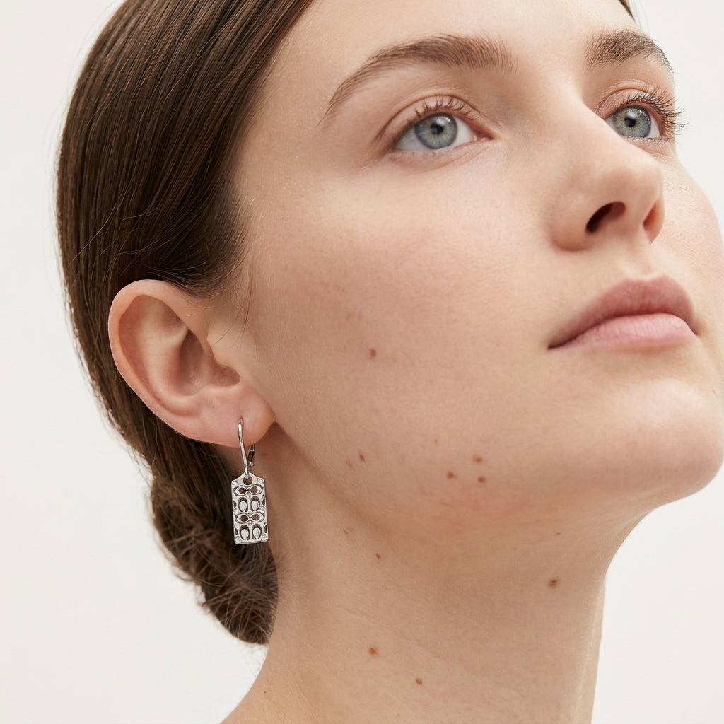 A close-up of a model wearing a silver dangling earring with an ornate filigree pattern, shown against a plain light background.