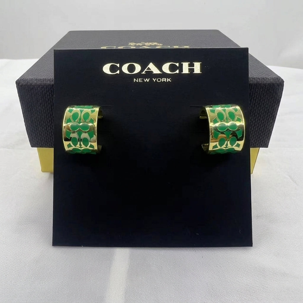 A pair of Coach hoop earrings with green and gold monogram pattern, displayed on a black Coach New York card holder in front of their black presentation box.