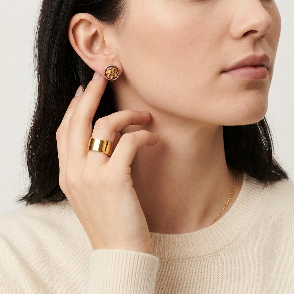 A close-up lifestyle shot of a model wearing a small gold stud earring with a circular gold-toned emblem, paired with a wide gold ring and a delicate gold chain necklace.