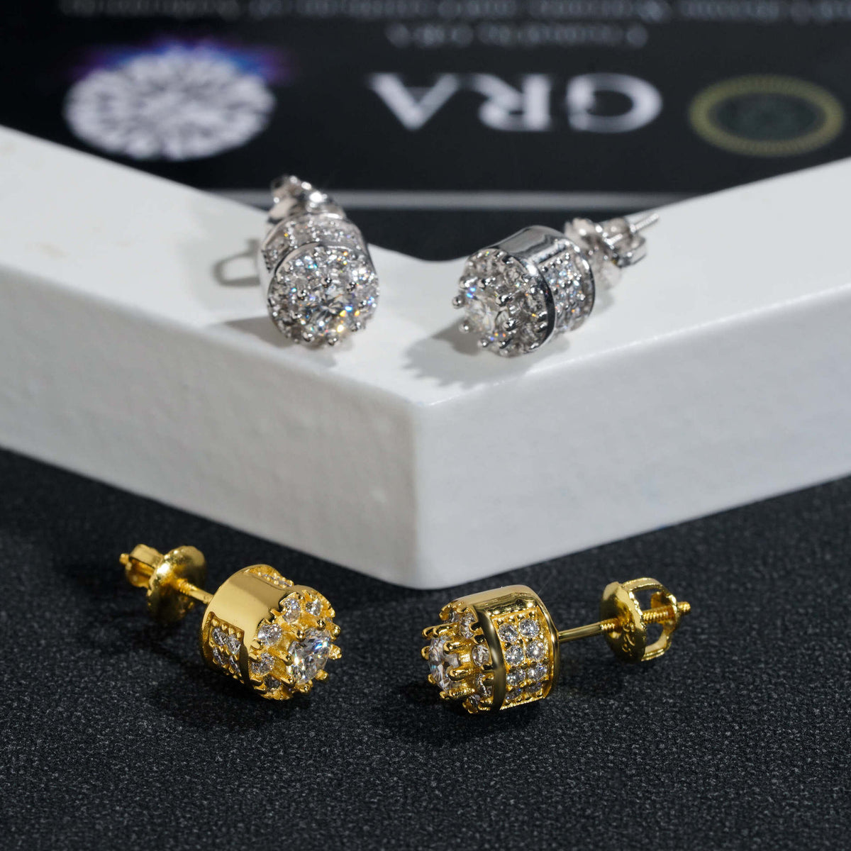 Pair of silver and yellow gold iced out 3D crown cylinder solitaire moissanite stud earrings displayed on white pedestal box