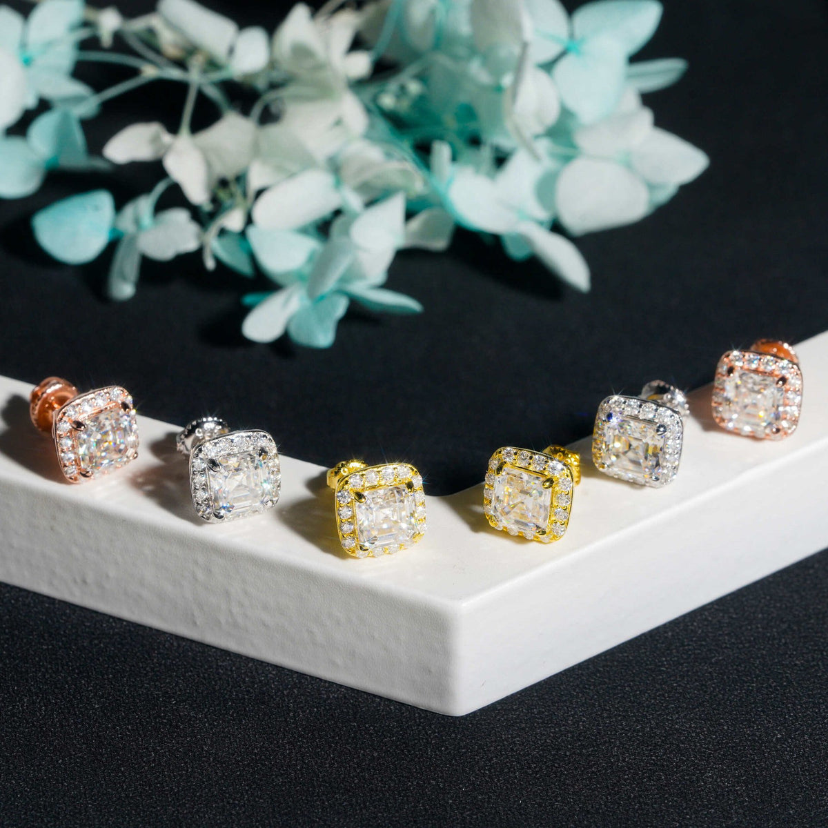 Rose gold, silver, and yellow gold iced out asscher cut halo solitaire moissanite stud earrings on white pedestal box   