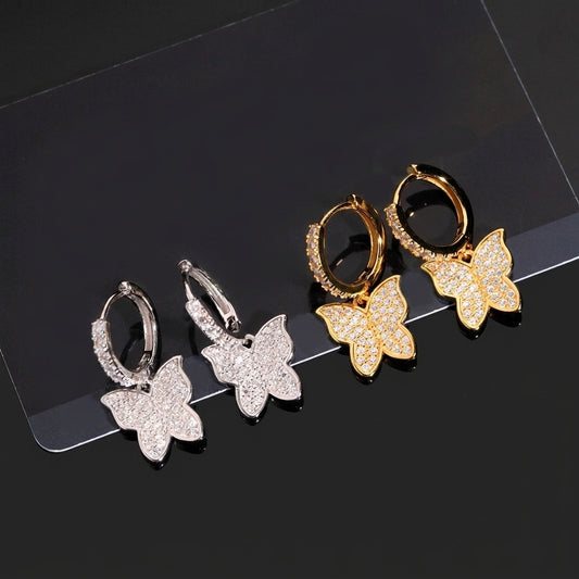Iced out butterfly dangle huggie hoop moissanite earrings in silver and yellow gold on dark surface