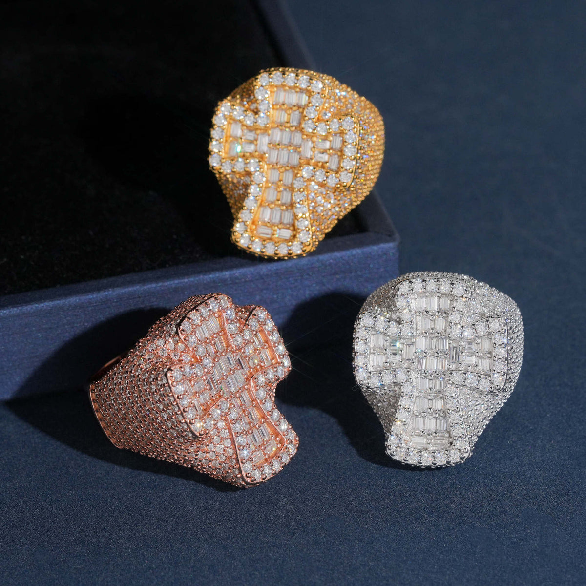Three tone display of iced out cross moissanite baguette statement rings resting on dark surface