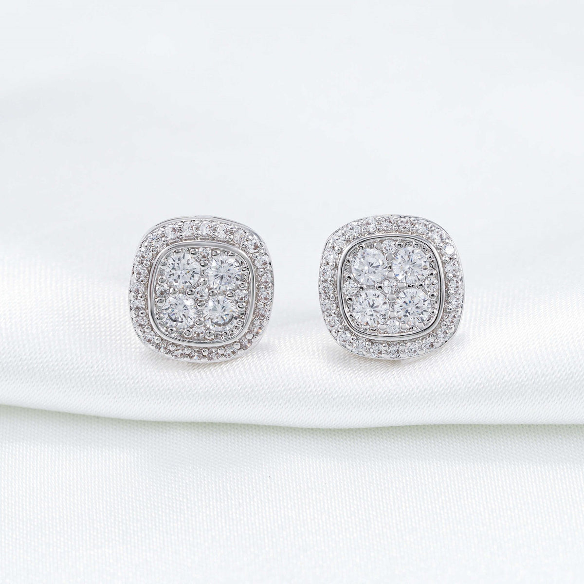 Pair of silver iced out cushion square cluster moissanite stud earrings on white silk cloth backdrop