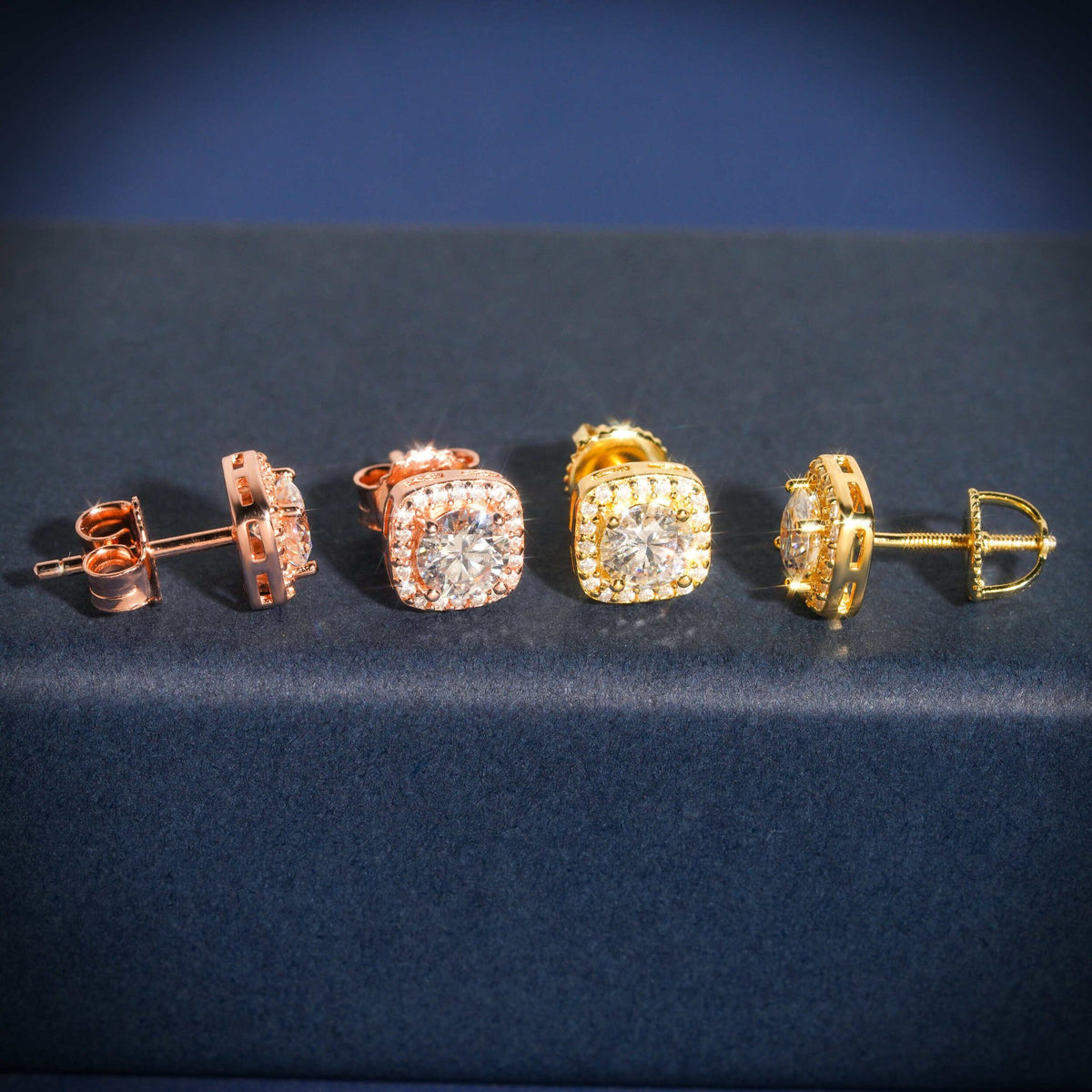 Rose gold and yellow gold iced out cushion square halo solitaire moissanite stud earrings aligned on navy display stand