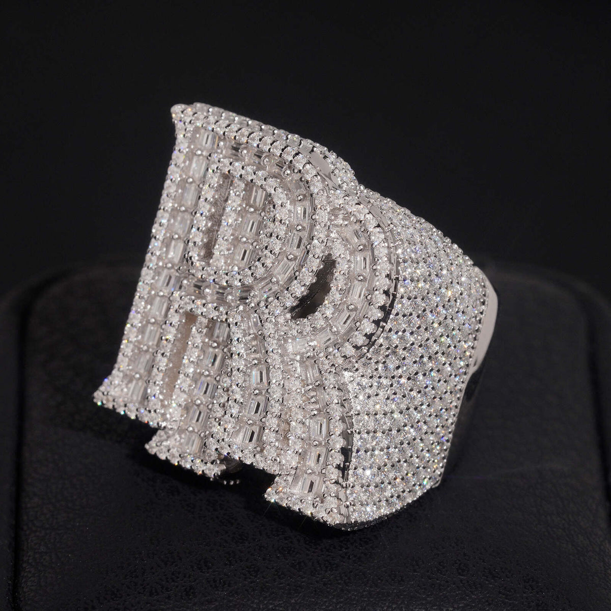 Angled view of iced out double letter RR moissanite baguette ring resting on dark surface