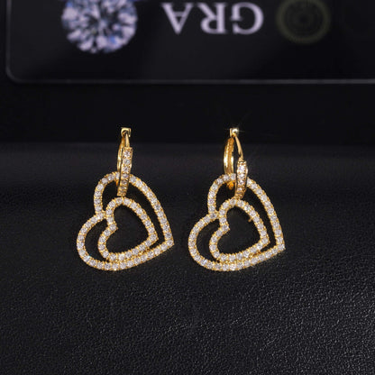 Pair of yellow gold iced out double open heart dangle huggie hoop moissanite earrings in front of GRA card