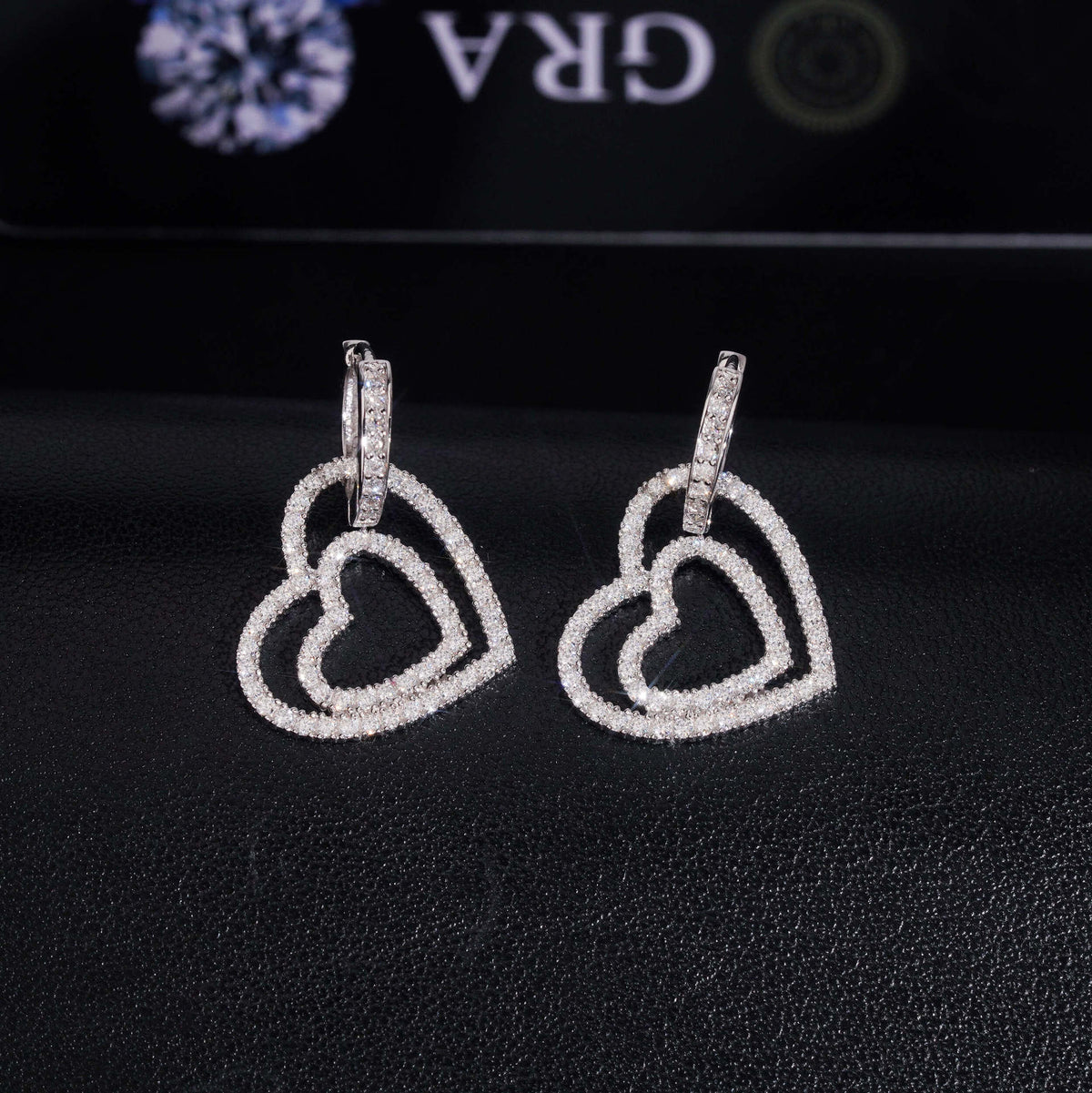 Pair of silver iced out double open heart dangle huggie hoop moissanite earrings displayed on GRA report card   