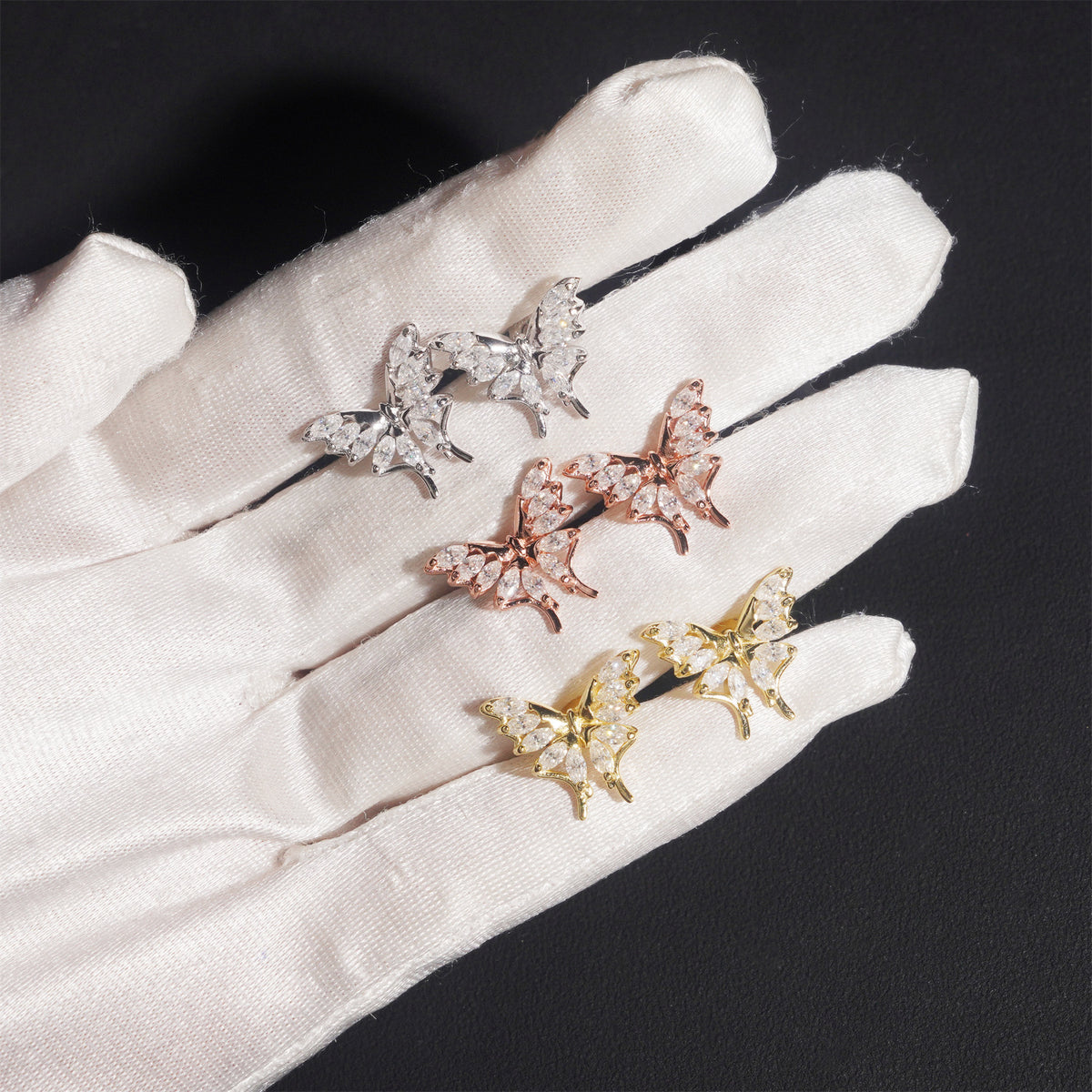 Gloved hand holding silver, rose gold, and yellow gold iced out marquise butterfly moissanite stud earrings for scale   