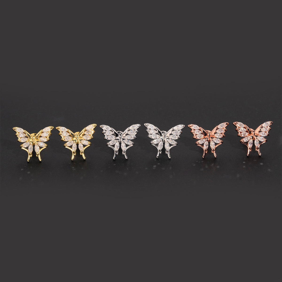 Iced out marquise butterfly moissanite stud earrings in yellow gold, silver, and rose gold on dark surface